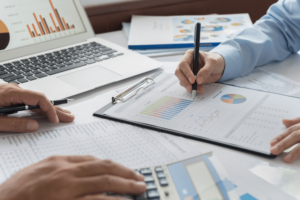 Two peoples hands hovering over accounting & finance documents on a desk with a laptop